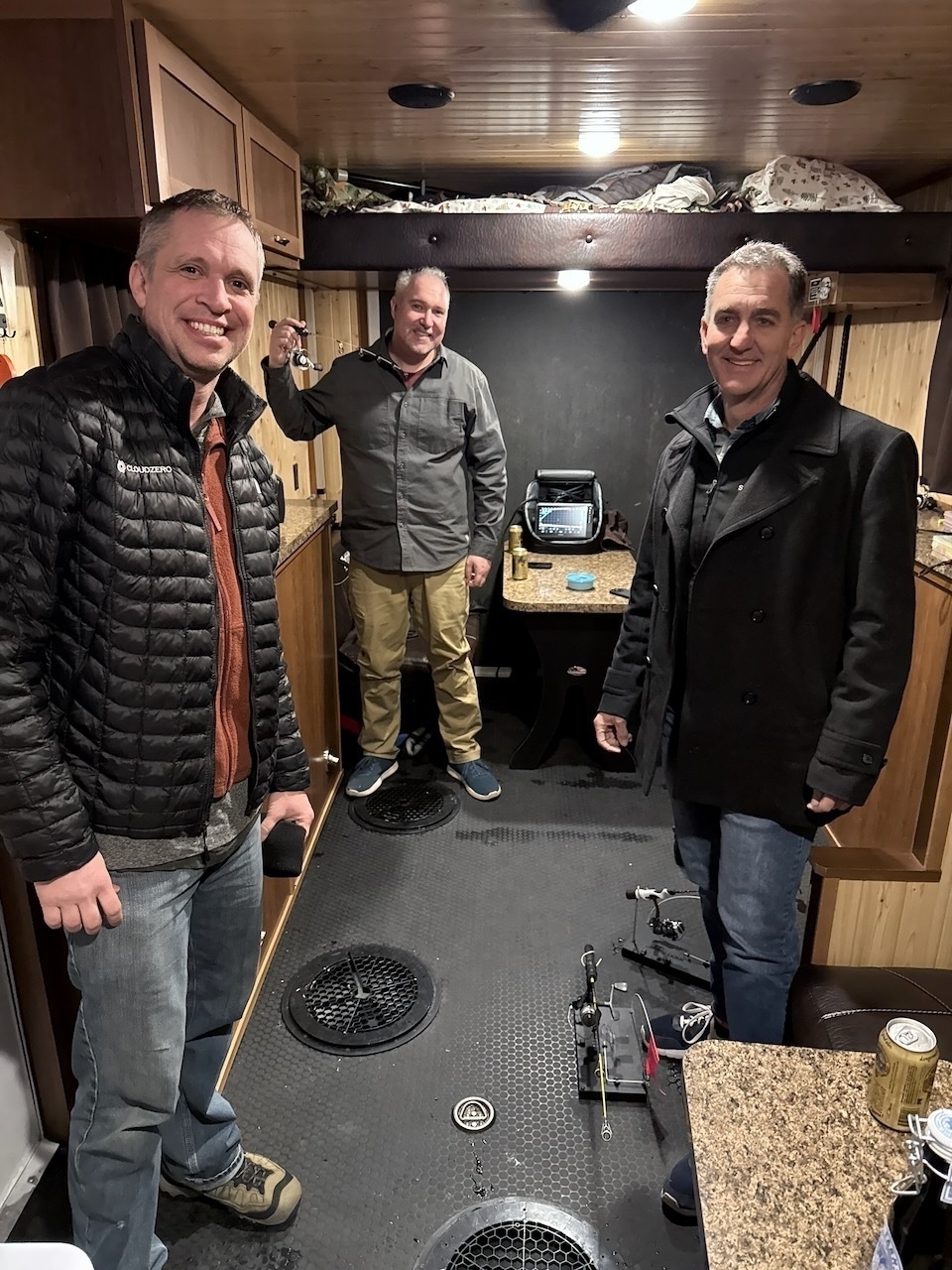 Three men are standing and smiling inside a cozy, wood-paneled room with some electronic equipment and a can on a counter.