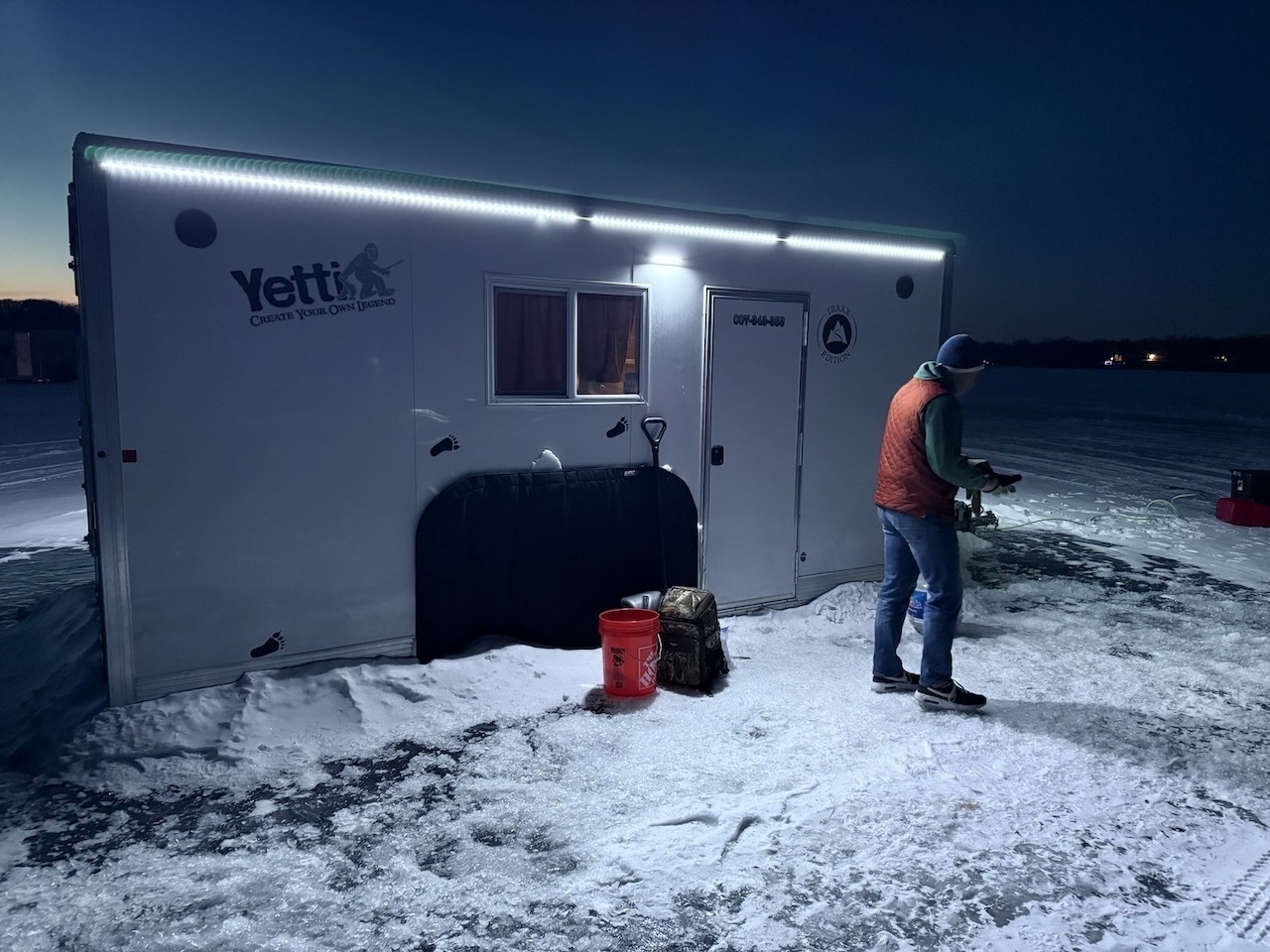 A person stands outside a small, brightly lit Yetti trailer on a snowy landscape at dusk.