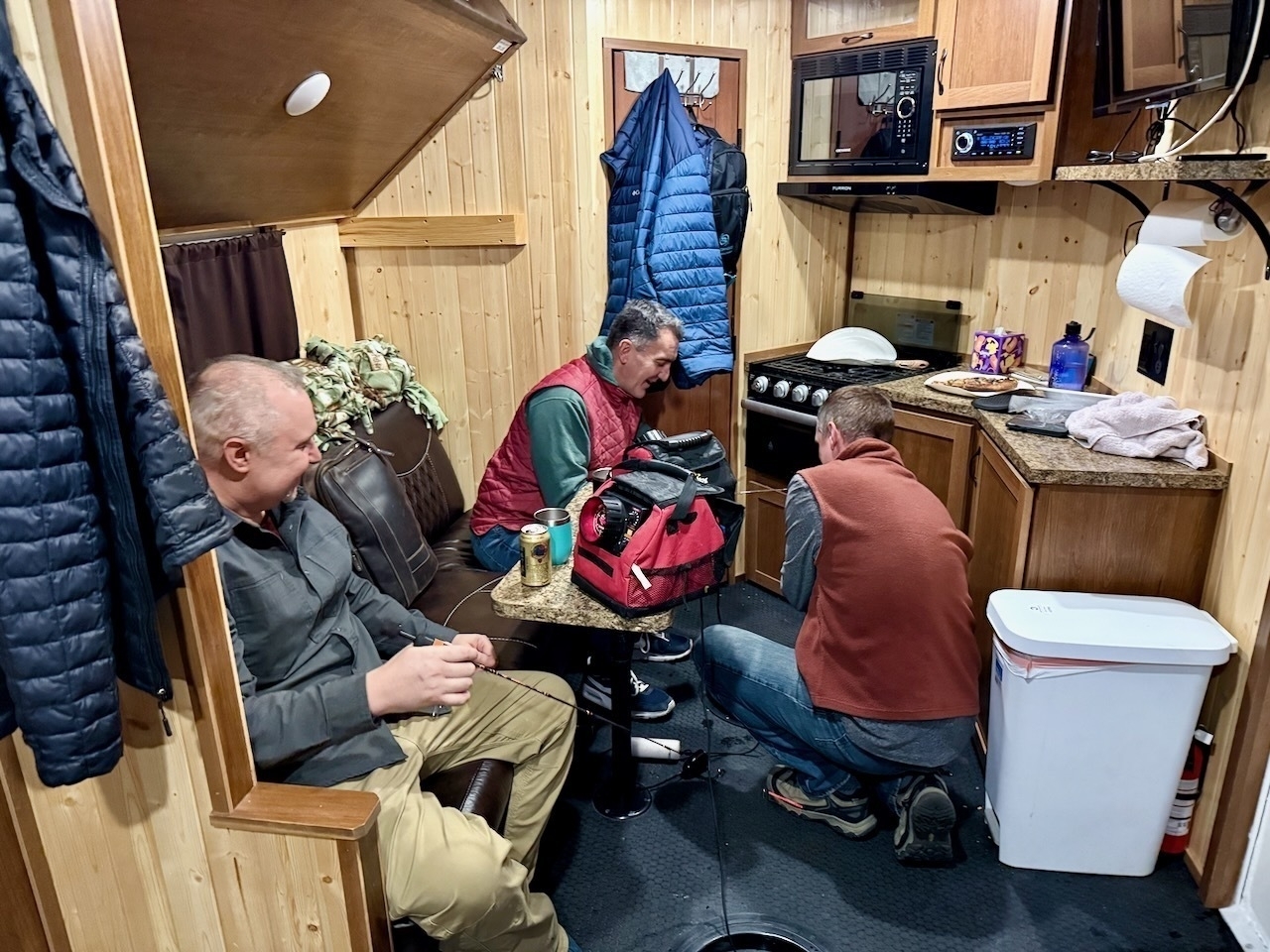 Three people are gathered inside a cozy wooden cabin, sitting and engaging in conversation, with clothing and kitchen amenities visible around them.