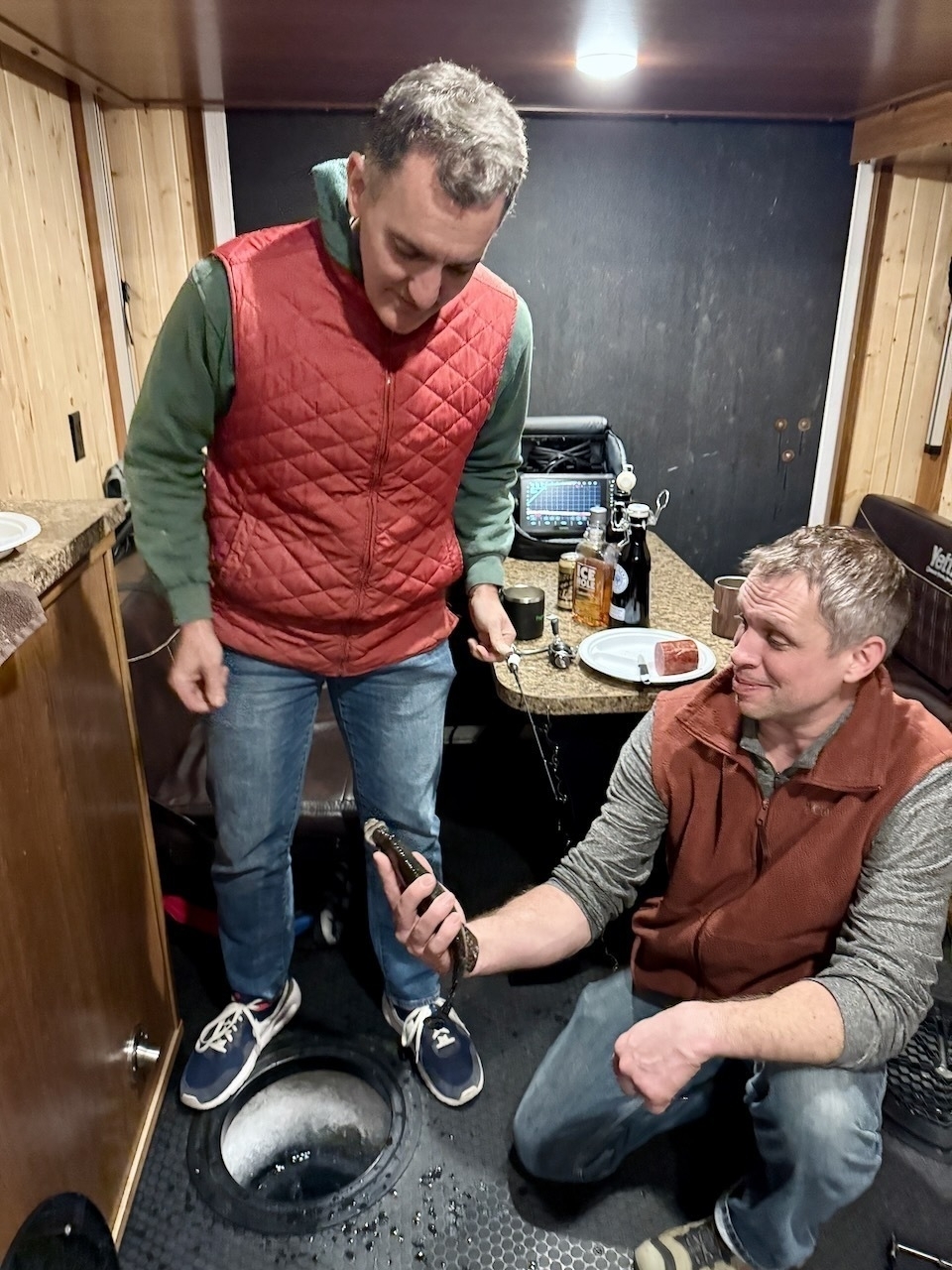 Two men inside a wooden ice fishing cabin are examining a fish by an ice hole, with a plate of food and a drink on the counter behind them.