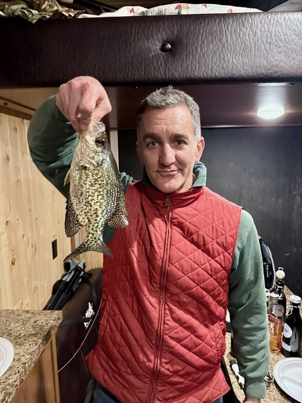 A person holding up a fish in a cozy indoor setting, wearing a red quilted vest and green hoodie.