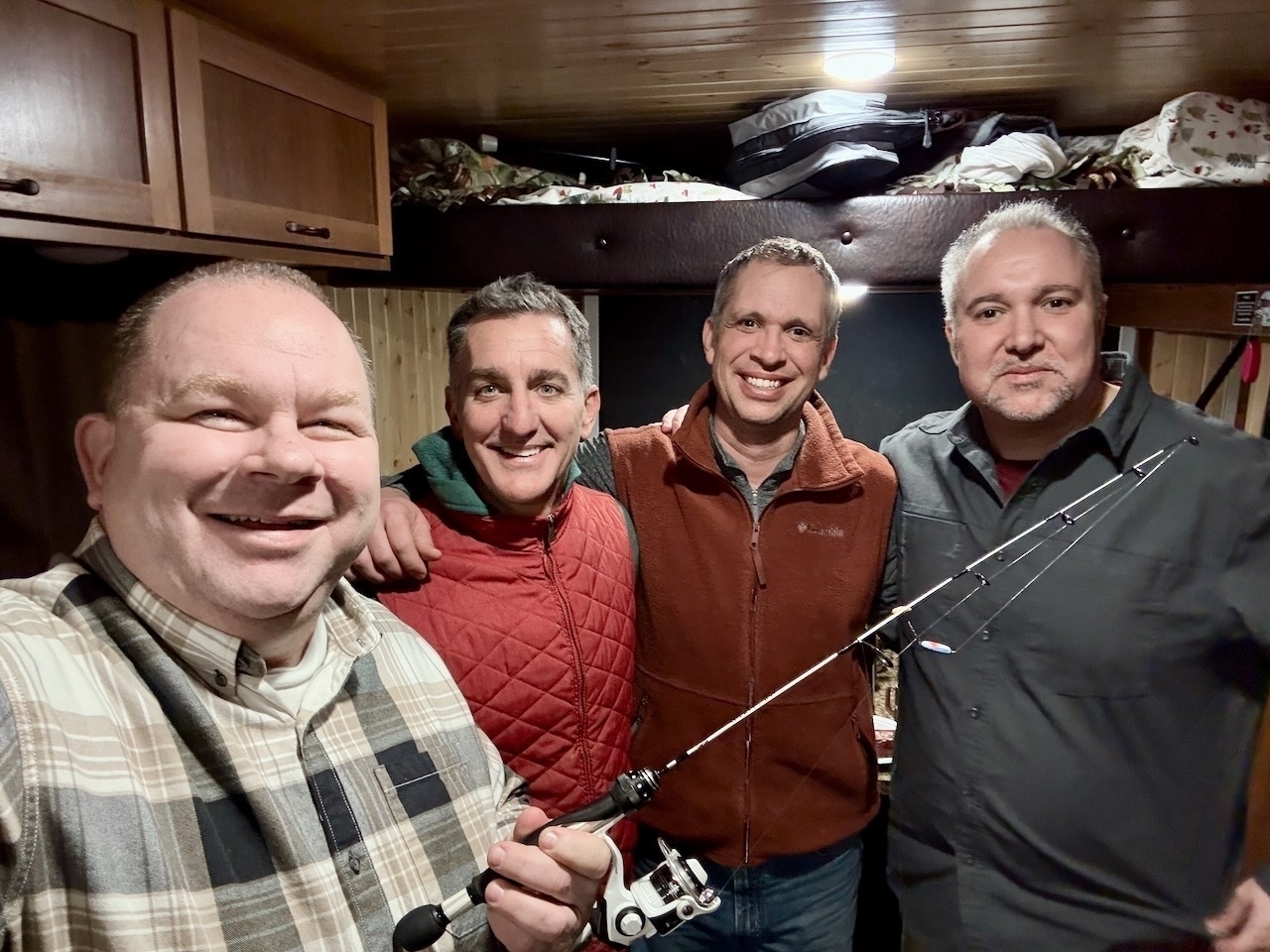 Four men posing together indoors, with one holding a fishing rod.